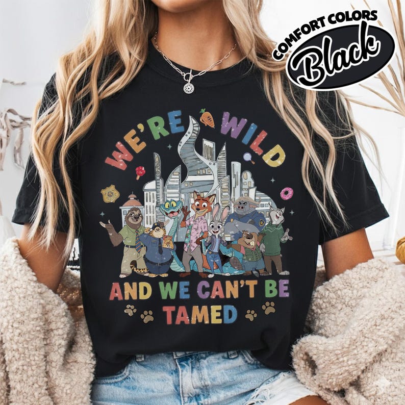 May include: Black t-shirt with a colorful graphic featuring characters from a cartoon movie. The text "WE'RE WILD AND WE CAN'T BE TAMED" is displayed in rainbow colors. The shirt also has the words "COMFORT COLORS" and "Black" in a circular design.