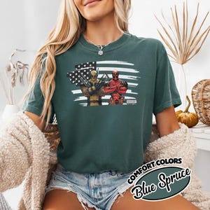May include: A dark green short-sleeve t-shirt featuring a graphic print of Wolverine and Deadpool standing in front of a stylized American flag. The design includes the text Parental Advisory in the bottom right corner of the graphic print.
