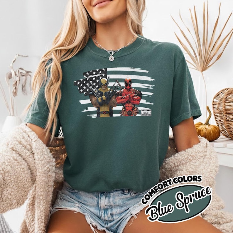 May include: A dark green short-sleeve t-shirt featuring a graphic print of Wolverine and Deadpool standing in front of a stylized American flag. The design includes the text Parental Advisory in the bottom right corner of the graphic print.