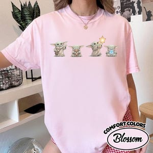 以下が含まれることがあります： 4つのGroguのイラストが描かれた、ライトピンクのComfort Colors Tシャツ。Groguのキャラクターは、おもちゃ、星、光るオブジェクトを持っています。シャツには、白い楕円の中に「Comfort Colors」と「Blossom」の文字が入っています。
