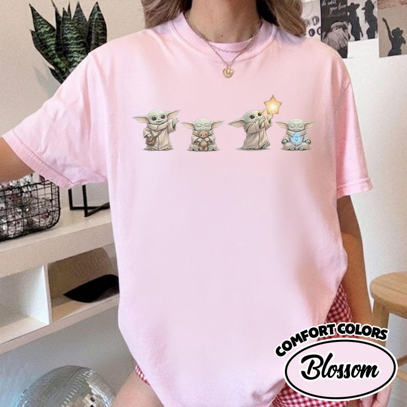 以下が含まれることがあります： 4つのGroguのイラストが描かれた、ライトピンクのComfort Colors Tシャツ。Groguのキャラクターは、おもちゃ、星、光るオブジェクトを持っています。シャツには、白い楕円の中に「Comfort Colors」と「Blossom」の文字が入っています。