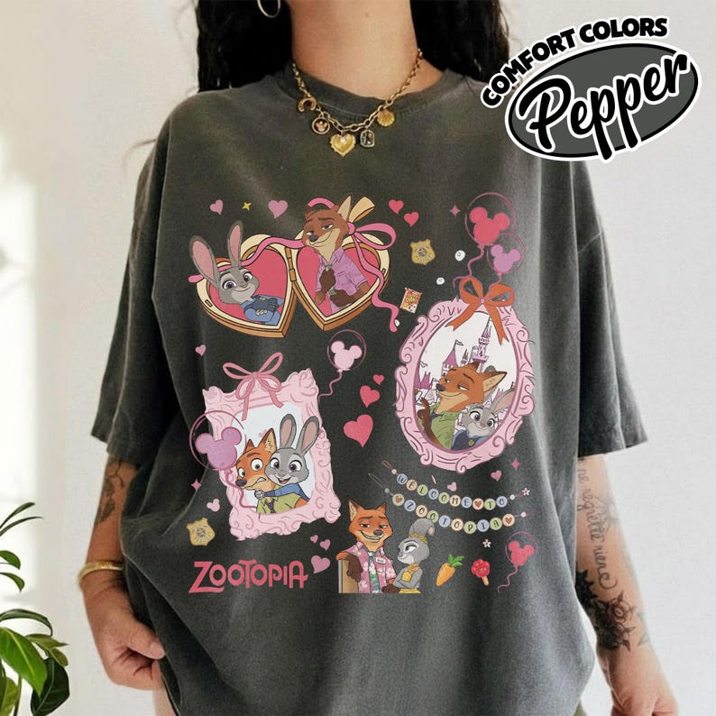 May include: A charcoal gray short-sleeve t-shirt featuring a graphic print of Zootopia characters Judy Hopps and Nick Wilde. The design includes heart-shaped and ornate frames, pink bows, small hearts, and the text Zootopia in a pink font.
