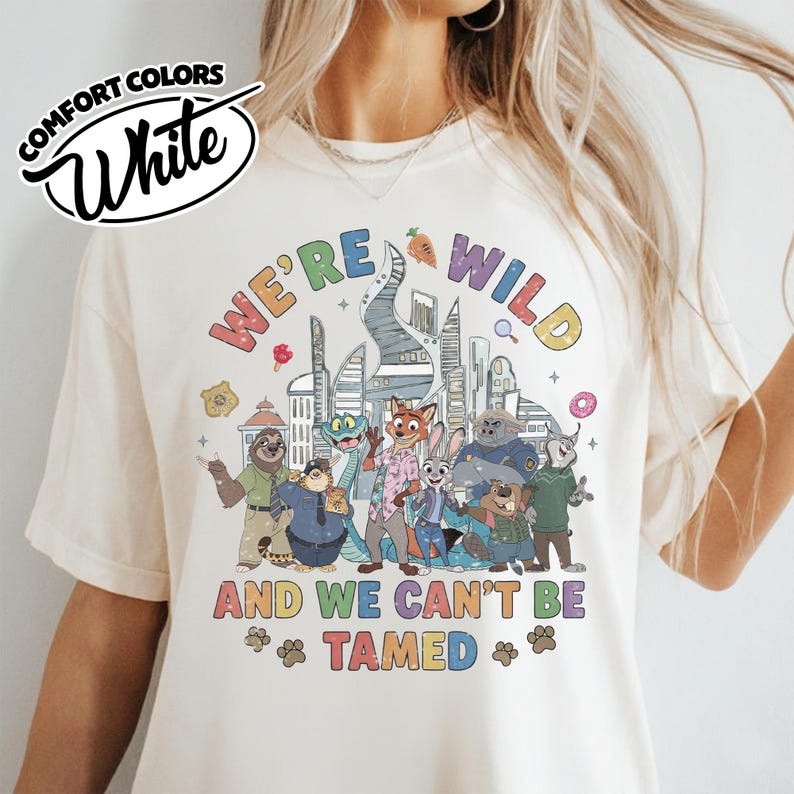May include: White Comfort Colors t-shirt featuring a colorful graphic of animated animal characters with the text "WE'RE WILD AND WE CAN'T BE TAMED". The design includes a cityscape and various food items.