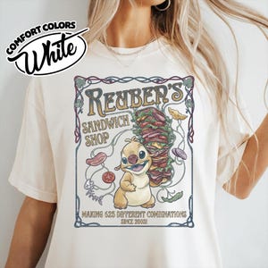 K&ouml;nnte beinhalten: Wei&szlig;es Comfort Colors T-Shirt mit einem Aufdruck f&uuml;r Reuben's Sandwich Shop. Das Design zeigt eine Cartoonfigur, die ein gro&szlig;es Sandwich h&auml;lt, mit dem Text "MAKING 625 DIFFERENT COMBINATIONS SINCE 2003!"