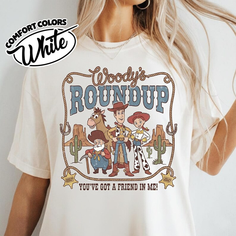 May include: White Comfort Colors t-shirt featuring a graphic of Woody's Roundup characters. The design includes Woody, Jessie, and other characters with the text "Woody's Roundup" and "You've Got a Friend in Me!"