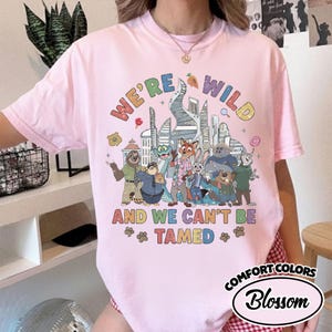 May include: A light pink Comfort Colors t-shirt with the text "WE'RE WILD AND WE CAN'T BE TAMED" and a colorful graphic of animal characters in front of a city skyline. The shirt has a crew neck and short sleeves.
