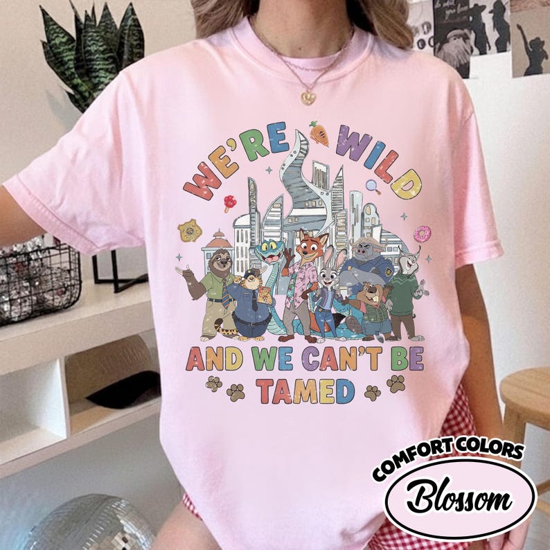 May include: A light pink Comfort Colors t-shirt with the text "WE'RE WILD AND WE CAN'T BE TAMED" and a colorful graphic of animal characters in front of a city skyline. The shirt has a crew neck and short sleeves.