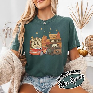 May include: Blue Spruce t-shirt featuring a graphic of Lightning McQueen and Mater from the movie Cars, with a Route 66 sign and Cozy Cone Motel. The shirt has the words "Comfort Colors" and "Blue Spruce" in a circular design.