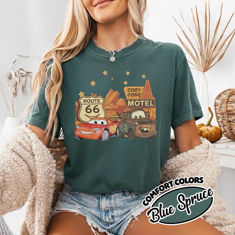 May include: Blue Spruce t-shirt featuring a graphic of Lightning McQueen and Mater from the movie Cars, with a Route 66 sign and Cozy Cone Motel. The shirt has the words "Comfort Colors" and "Blue Spruce" in a circular design.