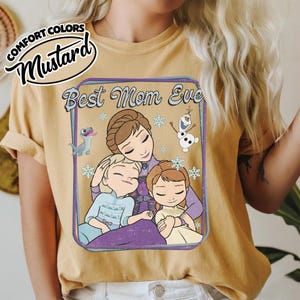 May include: Mustard-colored t-shirt featuring a graphic of a mother and two children with snowflakes and the text "Best Mom Ever." The graphic includes cartoon characters. The shirt has rolled-up sleeves and a Comfort Colors label.