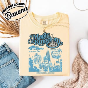 May include: A light yellow t-shirt with a graphic design featuring the words "Magic Kingdom" in a retro font, along with illustrations of Cinderella Castle, Autopia Car, Peter Pan's Flight Vehicle, and Tiki Room Drummers. The shirt also has the text "Comfort Colors" and "Banana".