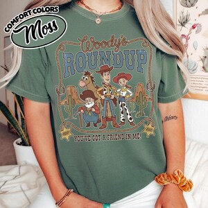 May include: Moss green t-shirt featuring a graphic of Woody's Roundup characters. The design includes Woody, Jessie, and other characters from the movie, with the text "Woody's Roundup" and "You've Got a Friend in Me!"