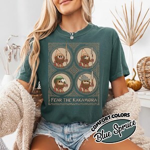 May include: Teal t-shirt with a graphic of four cartoon characters holding spears, each wearing a different hat. The text "FEAR THE KAKAMORA" is below the graphic. The shirt also has the text "COMFORT COLORS Blue Spruce".