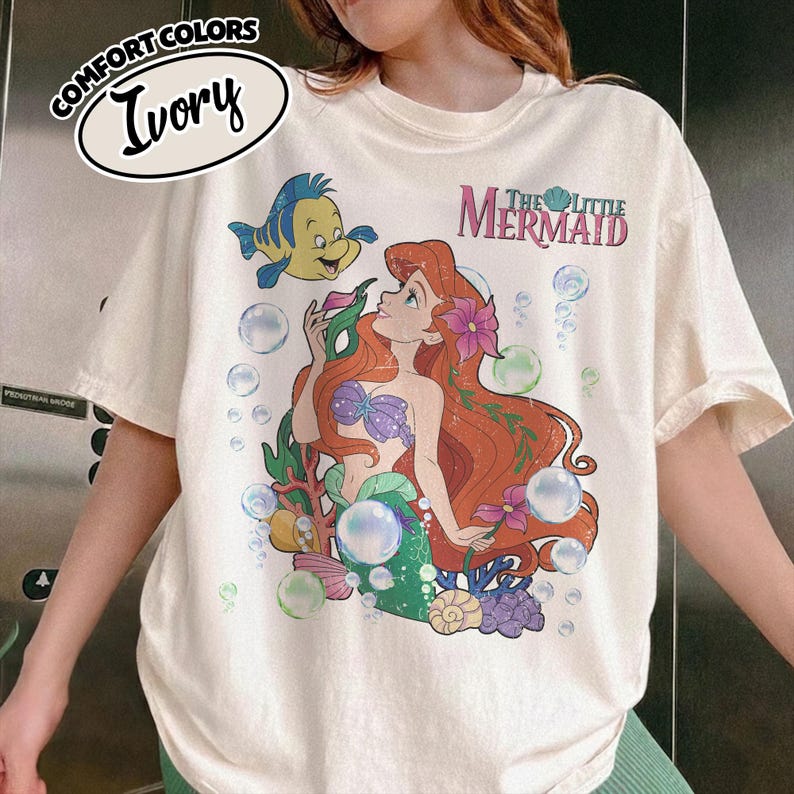 K&ouml;nnte beinhalten: Ein elfenbeinfarbenes T-Shirt mit einer Grafik von Arielle, die Meerjungfrau, mit rotem Haar, einem violetten Muschel-Oberteil und einem gr&uuml;nen Schwanz. Das Design zeigt Fabius, Blasen und den Text The Little Mermaid. Comfort Colors Ivory Etikett.