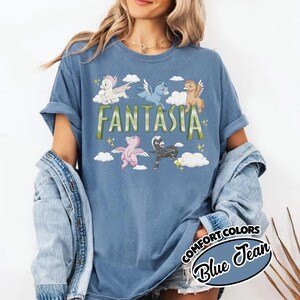 May include: A blue t-shirt with the word "FANTASIA" in green and gold lettering. The shirt features cartoon pegasus characters in various colours, including white, blue, brown, pink, and black, with clouds and stars. The shirt is labelled "Comfort Colors Blue Jean."