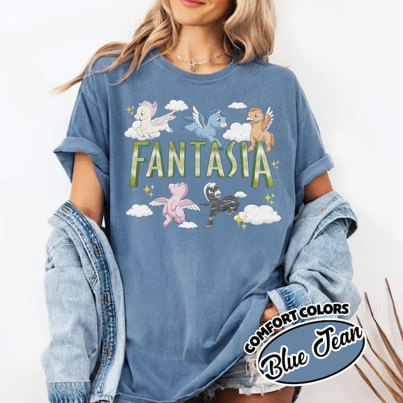 May include: A blue t-shirt with the word "FANTASIA" in green and gold lettering. The shirt features cartoon pegasus characters in various colours, including white, blue, brown, pink, and black, with clouds and stars. The shirt is labelled "Comfort Colors Blue Jean."