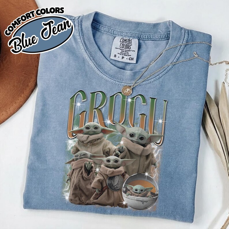 K&ouml;nnte beinhalten: Ein blaues T-Shirt in Jeansoptik mit einem Grafikdruck von Grogu in verschiedenen Posen. Der Text GROGU ist in einer gro&szlig;en, stilisierten Schrift &uuml;ber den Figuren gedruckt. Das T-Shirt hat ein Markenetikett von Comfort Colors am Kragen.