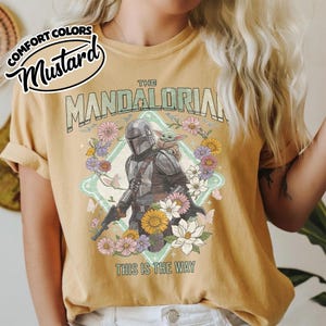 May include: A mustard yellow t-shirt featuring a graphic print of The Mandalorian and Grogu surrounded by colorful flowers and butterflies. The text reads THE MANDALORIAN at the top and THIS IS THE WAY at the bottom. A Comfort Colors Mustard label is visible.
