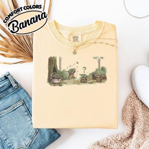 May include: A light yellow t-shirt with a graphic of Grogu playing in a forest setting. The shirt has the text "Comfort Colors Banana" in a black and white oval. The shirt is paired with blue jeans and a white shoe.