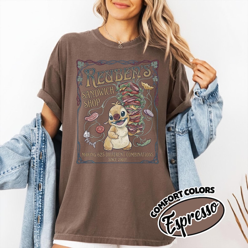 K&ouml;nnte beinhalten: Braunes T-Shirt mit einer Cartoonfigur und dem Text "Reuben's Sandwich Shop". Das Design umfasst einen Stapel Sandwiches und Lebensmittelillustrationen. Das Shirt hat auch den Text "Making 625 Different Combinations Since 2003."