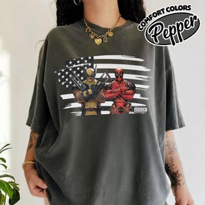 May include: A charcoal gray short-sleeve t-shirt featuring a graphic print of Wolverine and Deadpool standing in front of a stylized American flag. The shirt includes a small rectangular label with the text "ADVISORY" on the lower right side.
