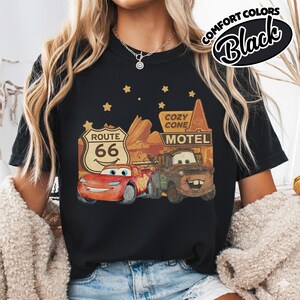 May include: Black t-shirt featuring a graphic of Lightning McQueen and Mater from the movie Cars, with Route 66 and Cozy Cone Motel signage. The shirt has the words "Comfort Colors" and "Black" in a circular design.