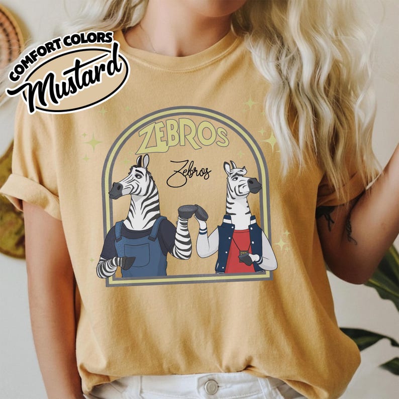 May include: Mustard-coloured t-shirt with a cartoon design of two zebras inside a yellow arch, with the text "ZEBROS" above and "Zebros" below. The shirt also has the text "COMFORT COLORS" and "Mustard" in a circular design.