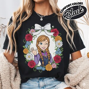 Peut inclure: T-shirt noir avec une illustration de dessin anim&eacute; d'un personnage aux cheveux tress&eacute;s, faisant un clin d'&oelig;il et saluant, entour&eacute; d'une couronne florale et d'un n&oelig;ud blanc. Les mots "COMFORT COLORS" et "Black" sont imprim&eacute;s en haut &agrave; droite.