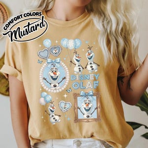 May include: Mustard-colored t-shirt featuring a Disney Olaf design. The design includes multiple images of Olaf the snowman, blue balloons, and the text "Disney Olaf". The shirt has a relaxed fit and short sleeves.