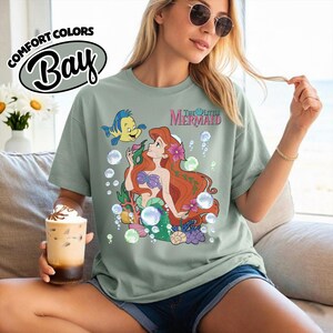 K&ouml;nnte beinhalten: Ein kurz&auml;rmeliges T-Shirt in Salbeigr&uuml;n mit einem Grafikdruck von Arielle und Fabius aus Arielle, die Meerjungfrau, umgeben von Blasen, Korallen und Blumen. Der Text The Little Mermaid ist in Pink &uuml;ber den Figuren auf der Vorderseite gedruckt.