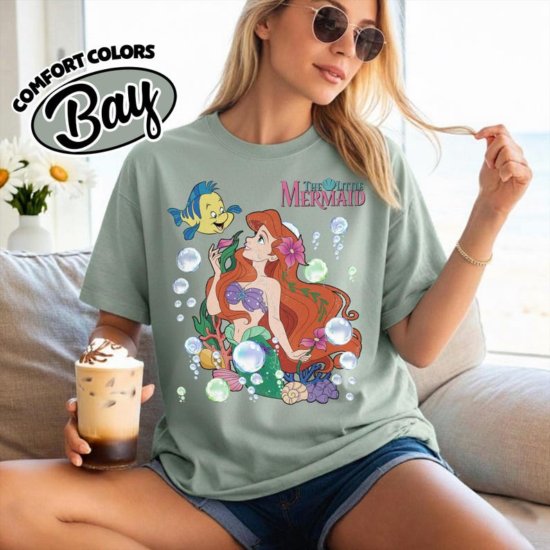 K&ouml;nnte beinhalten: Ein kurz&auml;rmeliges T-Shirt in Salbeigr&uuml;n mit einem Grafikdruck von Arielle und Fabius aus Arielle, die Meerjungfrau, umgeben von Blasen, Korallen und Blumen. Der Text The Little Mermaid ist in Pink &uuml;ber den Figuren auf der Vorderseite gedruckt.