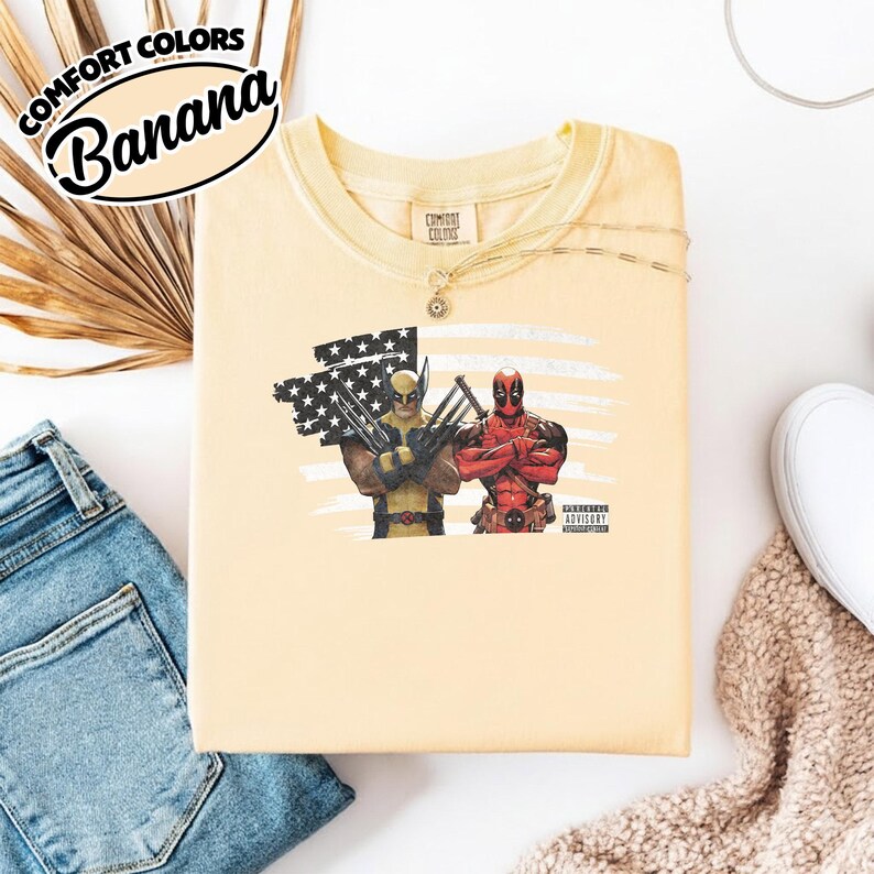 May include: A pale yellow t-shirt featuring a graphic of Wolverine and Deadpool standing in front of a stylized American flag. The shirt has a crew neckline and a Comfort Colors label at the inner collar. A small Parental Advisory logo is visible.