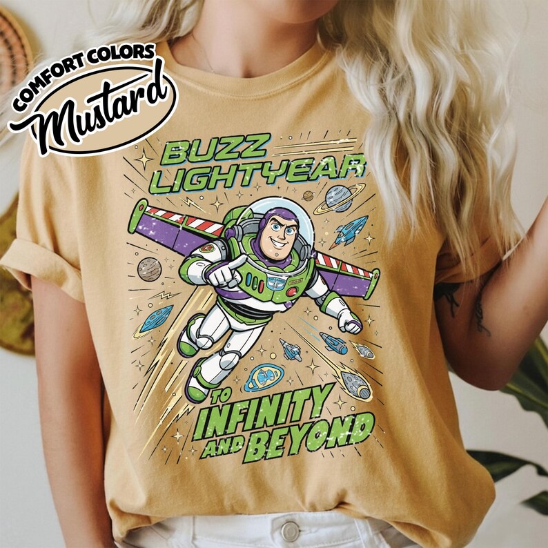 May include: A mustard yellow t-shirt featuring a graphic of Buzz Lightyear flying through space. The text reads BUZZ LIGHTYEAR and TO INFINITY AND BEYOND. The design includes planets, stars, and small spacecraft surrounding the character.