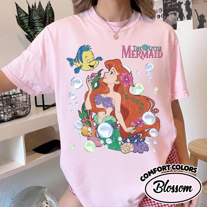 K&ouml;nnte beinhalten: Ein hellrosa T-Shirt mit einem Grafikdruck von Arielle und Fabius aus Arielle, die Meerjungfrau, umgeben von Blasen, Seetang und Muscheln. Der Text The Little Mermaid ist in Rosa und T&uuml;rkis &uuml;ber den Figuren aufgedruckt.