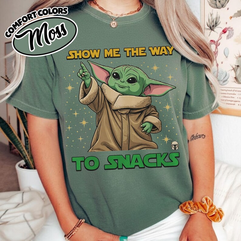 May include: Moss green t-shirt featuring a graphic of Grogu from The Mandalorian, pointing upwards. The text reads "SHOW ME THE WAY TO SNACKS" in yellow and green. The shirt also has the words "COMFORT COLORS" and "Moss" in a circular design.