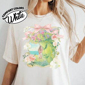 K&ouml;nnte beinhalten: Wei&szlig;es Comfort Colors T-Shirt mit einer Aquarell-Illustration. Das Design zeigt ein Frauengesicht mit Blumenkrone, einer rosa Schleife und einer Strandszene. Der Text "White" ist in Schreibschrift.