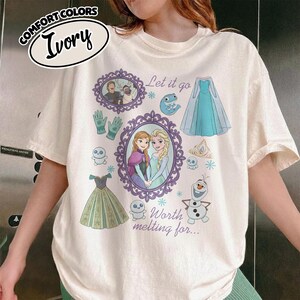 Op de afbeelding: Ivoor kleurig T-shirt met een grafisch ontwerp met personages en elementen uit een populaire animatiefilm. Het ontwerp bevat ingelijste portretten, jurken, handschoenen en de zinnen "Let it go" en "Worth melting for..."