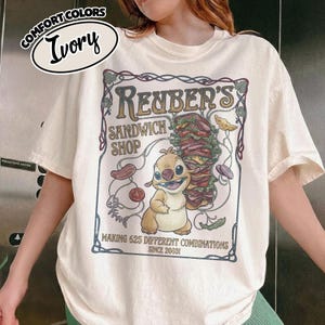 K&ouml;nnte beinhalten: Elfenbeinfarbenes T-Shirt mit einem Cartoon-Motiv f&uuml;r Reuben's Sandwich Shop. Das Design umfasst eine Zeichentrickfigur, ein gro&szlig;es Sandwich und den Text "MAKING 625 DIFFERENT COMBINATIONS SINCE 2003!". Das Shirt hat die Worte "COMFORT COLORS" und "Ivory" in einem Kreis.