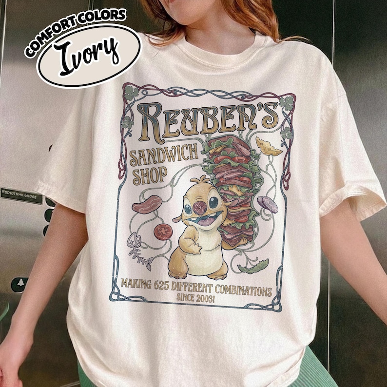 K&ouml;nnte beinhalten: Elfenbeinfarbenes T-Shirt mit einem Cartoon-Motiv f&uuml;r Reuben's Sandwich Shop. Das Design umfasst eine Zeichentrickfigur, ein gro&szlig;es Sandwich und den Text "MAKING 625 DIFFERENT COMBINATIONS SINCE 2003!". Das Shirt hat die Worte "COMFORT COLORS" und "Ivory" in einem Kreis.