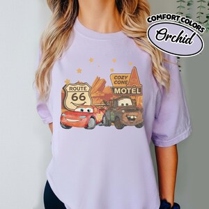 May include: Orchid Comfort Colors t-shirt featuring a graphic of Lightning McQueen and Mater from the movie Cars, with Route 66 and Cozy Cone Motel signs. The design is set against a desert backdrop with star accents.