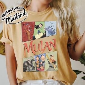 Puede incluir: Camiseta de color mostaza con la palabra "Mulan" en naranja y un gr&aacute;fico de personajes de la pel&iacute;cula animada. La camiseta tiene un aspecto vintage y desgastado, con las palabras "Comfort Colors" y "Mustard" en un dise&ntilde;o circular.