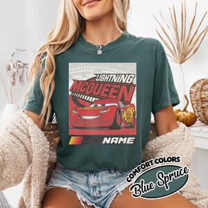 May include: A dark teal t-shirt featuring a graphic print of the red race car Lightning McQueen. The design includes the text Lightning McQueen above the car and the word NAME below it, set against a speedometer background with yellow accents.
