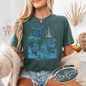 May include: Blue Spruce t-shirt with a graphic of the Magic Kingdom logo and attractions. The design includes Autopia Car, Peter Pan's Flight Vehicle, Tiki Room Drummer, and Cinderella Castle. The shirt is a Comfort Colors brand.