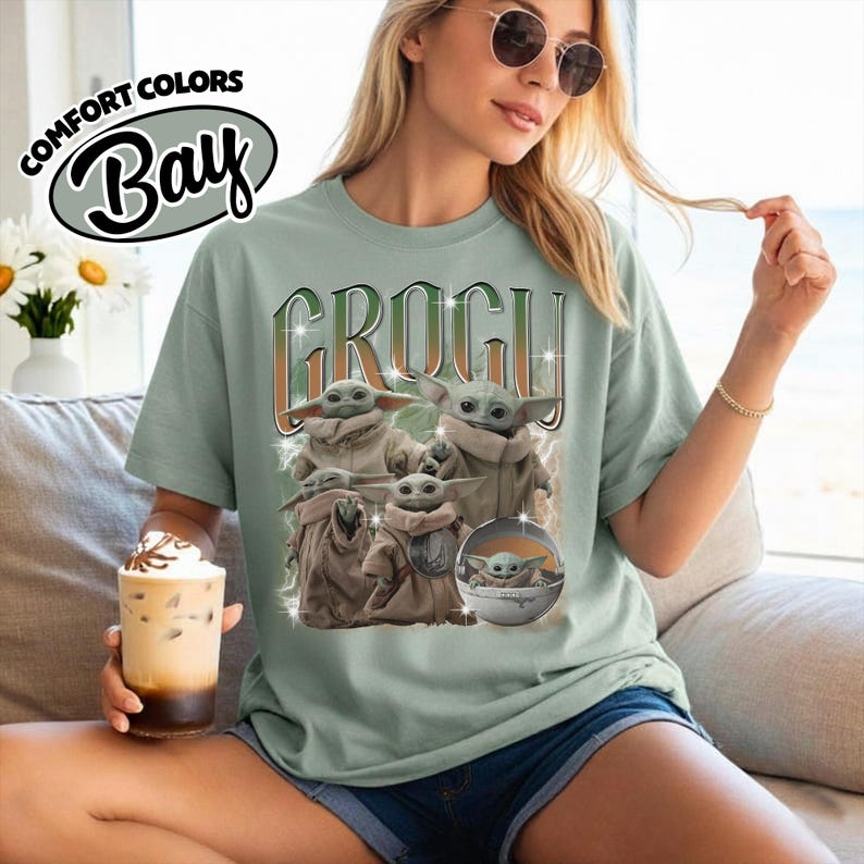 K&ouml;nnte beinhalten: Ein salbeigr&uuml;nes Kurzarm-T-Shirt mit dem Text COMFORT COLORS BAY und einem Grafikdruck von vier Grogu-Figuren aus The Mandalorian. Das zentrale Design zeigt den Namen GROGU in gro&szlig;en Buchstaben &uuml;ber den kleinen gr&uuml;nen Kreaturen.