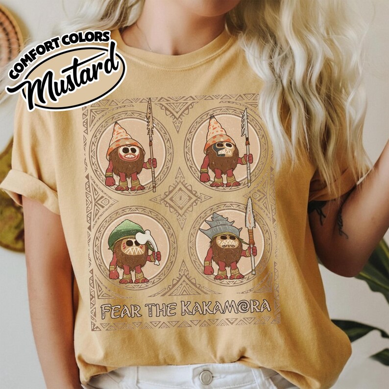 May include: Mustard-coloured t-shirt with a graphic of four cartoon figures with spears and hats, each in a circular frame. The text "FEAR THE KAKAMORA" is printed below the graphic. The shirt also has the text "COMFORT COLORS" and "Mustard" in a banner.