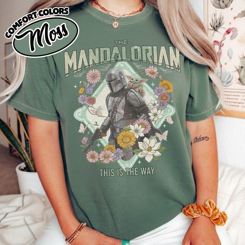 May include: A sage green short-sleeve t-shirt featuring a graphic of The Mandalorian and Grogu surrounded by colorful flowers and butterflies. The text reads THE MANDALORIAN at the top and THIS IS THE WAY at the bottom. A small oval logo reads COMFORT COLORS Moss.