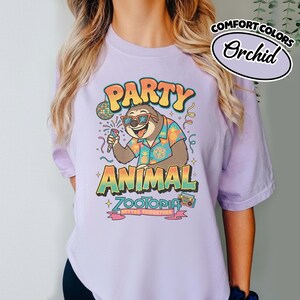Puede incluir: Camiseta Orchid Comfort Colors con un dise&ntilde;o de perezoso con gafas de sol y una camisa hawaiana, sosteniendo un micr&oacute;fono. La camiseta tiene las palabras "PARTY ANIMAL ZOOTOPIA" en texto colorido, con una bola de discoteca y una radio.