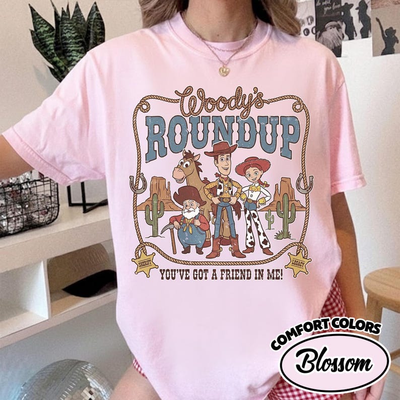 May include: A light pink t-shirt featuring a graphic of Woody's Roundup characters. The design includes Woody, Jessie, Bullseye, and other characters in a desert scene. Text includes "Woody's Roundup" and "You've Got a Friend in Me!"
