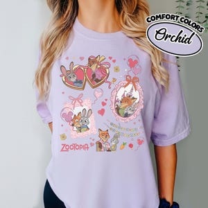 May include: A light purple t-shirt featuring a Zootopia graphic print with characters Judy Hopps and Nick Wilde. The design includes heart-shaped frames, balloons, a castle, and the text Zootopia. A label in the corner reads Comfort Colors Orchid.