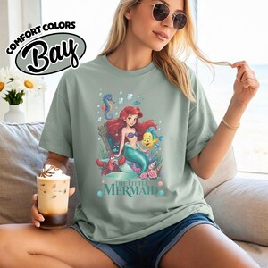 May include: A light green t-shirt featuring a colorful graphic of Ariel from The Little Mermaid. The design includes Ariel, Flounder, a seahorse, and the words "The Little Mermaid". The shirt also has the words "Comfort Colors Bay" printed on it.